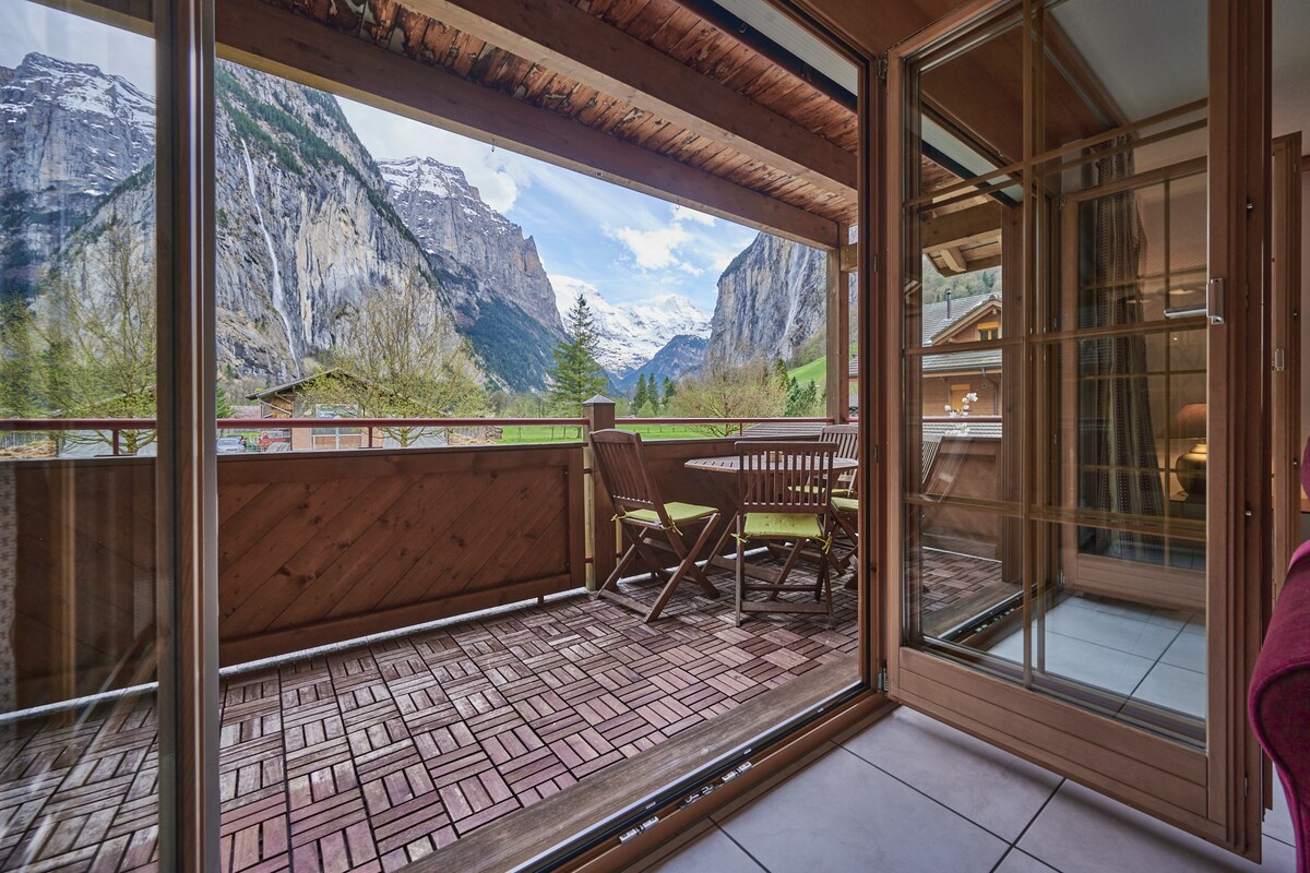 A spacious balcony is presented with wooden furniture and a table set for outdoor dining. The surrounding mountains are visible in the background, with striking cliffs framing the view. Glass doors open to the indoors, providing a seamless transition between spaces.