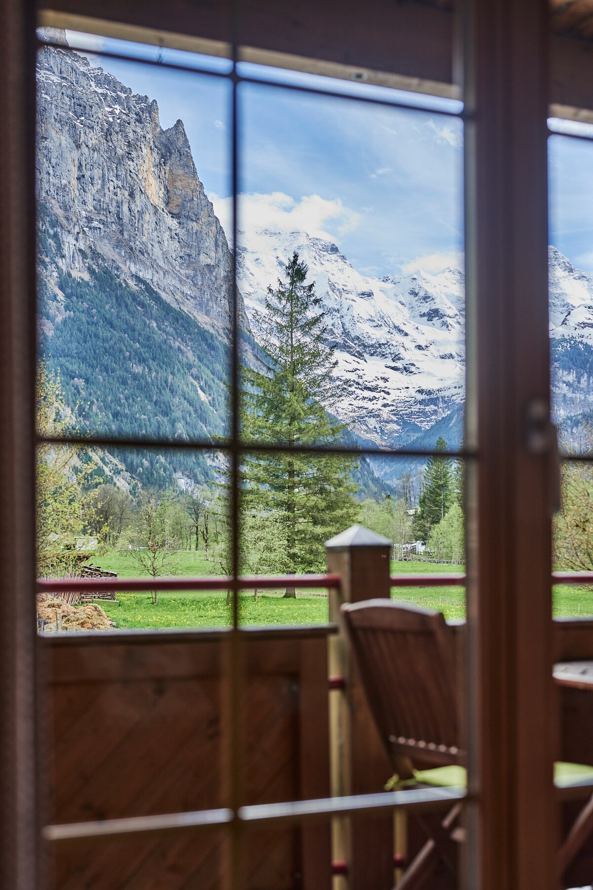 A panoramic view is visible from the balcony, showcasing snow-capped mountains and lush greenery. A wooden table and chairs are positioned on the balcony, framed by large windows that enhance the connection to the stunning outdoor landscape.