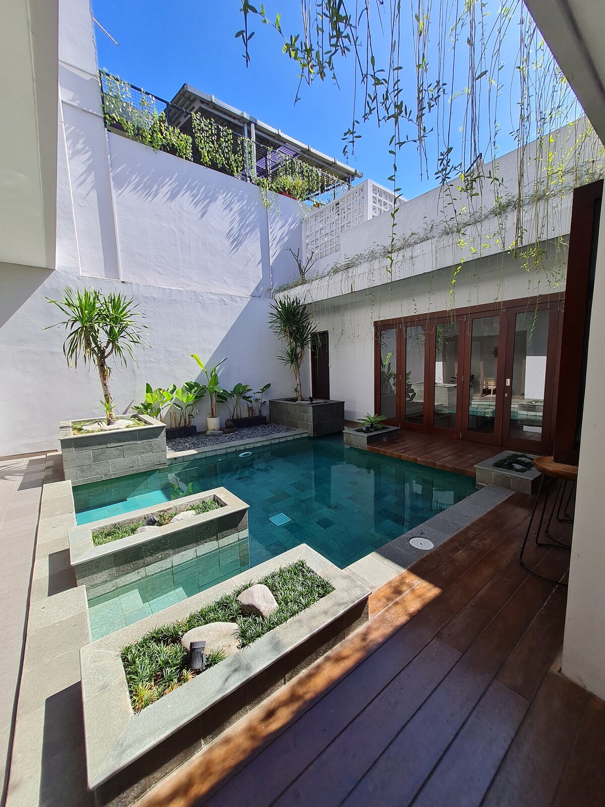 A serene private swimming pool is surrounded by lush greenery and modern stone planters. The pool is framed by large glass doors that open to the villa, allowing natural light to fill the space. Sunlight reflects off the calm water, creating a tranquil atmosphere.