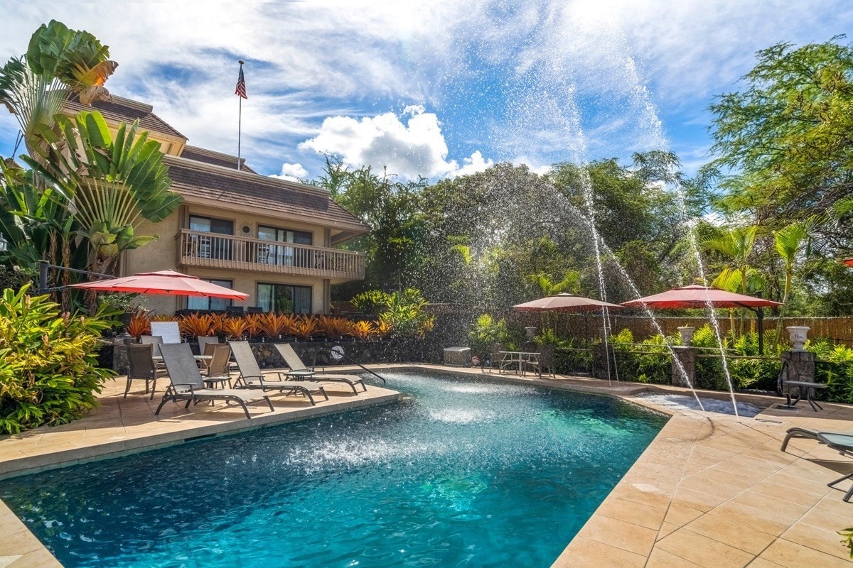 A tranquil pool area features a sparkling swimming pool surrounded by lounge chairs and tropical landscaping. Umbrellas provide shade for seating options, while water features create an inviting atmosphere. The two-story building with a balcony stands in the backdrop, framed by lush greenery under a clear sky.