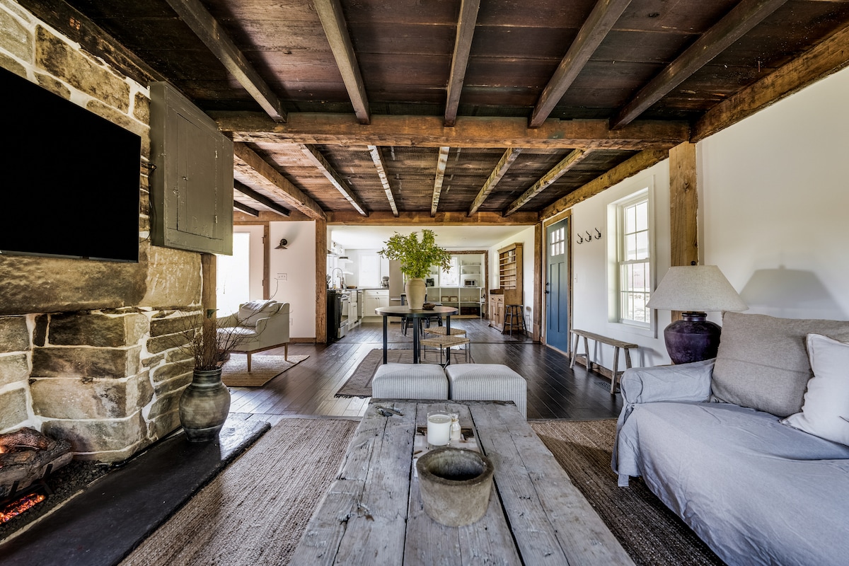 A spacious living area showcases exposed wooden beams and a stone fireplace. A cozy sofa and low coffee table are positioned on a textured rug. Natural light enters through multiple windows, illuminating the open space that leads to the kitchen.