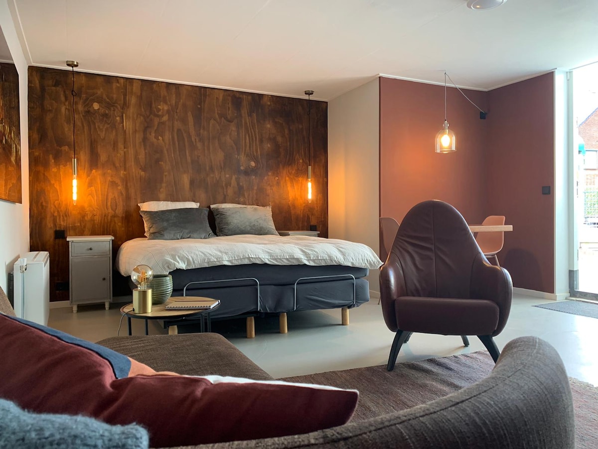 A stylish open space showcases a spacious bed against a warm wooden accent wall. A seating area is highlighted by a modern armchair, complemented by a cozy couch. Soft lighting from hanging fixtures adds a serene ambiance, with a small dining table visible in the background.