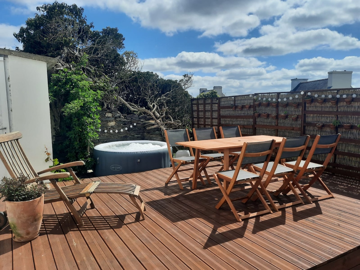 A wooden decking area features a table surrounded by six folding chairs, offering space for outdoor dining. A hot tub is positioned to one side, with greenery visible in the background. Bright clouds drift across a blue sky, creating a serene atmosphere.