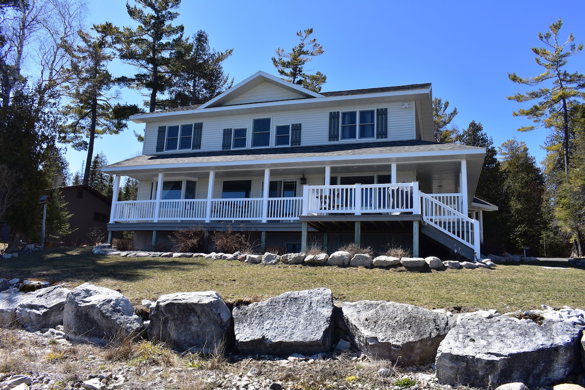 A lakeside house is presented, showcasing a large front porch with white railing. The two-story structure features multiple windows and is surrounded by a grassy area with rocks, with tall trees visible in the background against a clear blue sky.