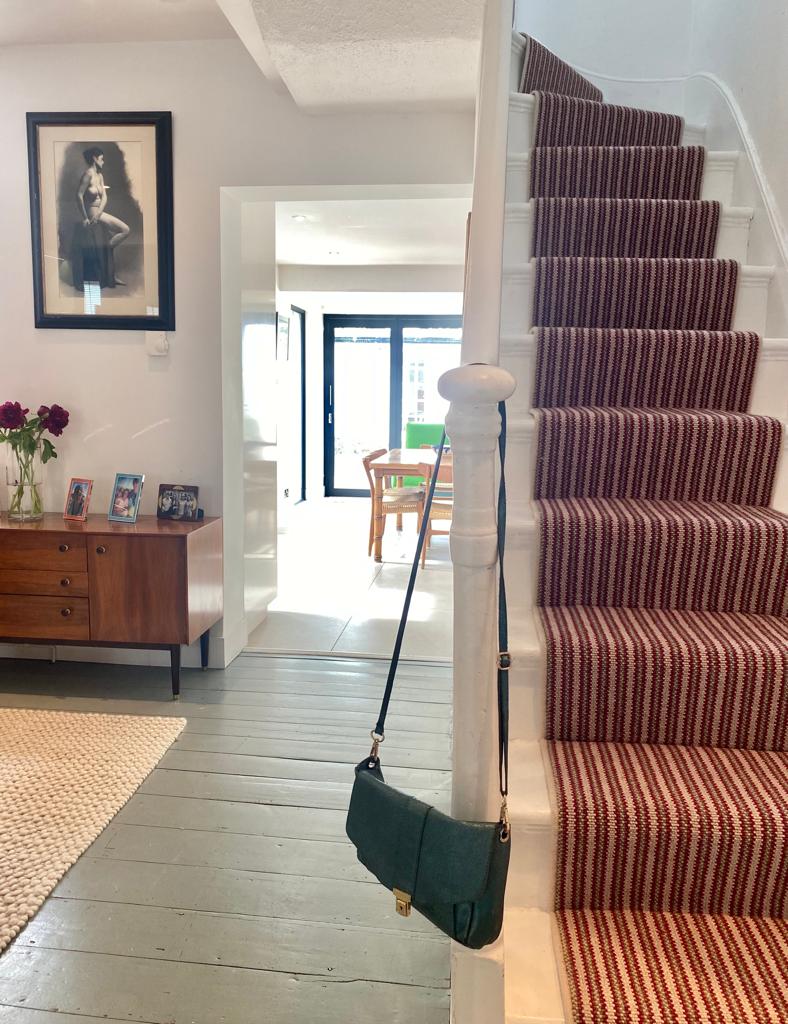 A staircase with red and white striped carpeting is featured, leading to an upper level. At the base, a wooden sideboard with framed photos and a vase of flowers is visible. A doorway leads to an open space illuminated by natural light, with glimpses of a dining area and greenery beyond.