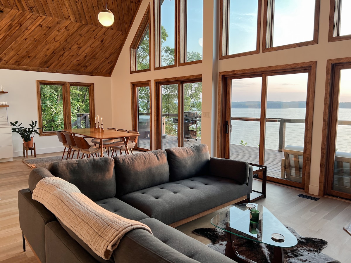 An open concept living area features a spacious gray sectional couch and large glass windows that offer views of Kentucky Lake. A wooden dining table is visible in the background, surrounded by chairs. Natural light fills the room, enhancing the wooden ceiling and warm tones.