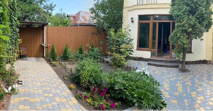 A private yard is surrounded by greenery and flowers, featuring a well-maintained garden path made of stone. The outdoor space includes a seating area and a wooden structure at one end, providing a serene environment for relaxation.