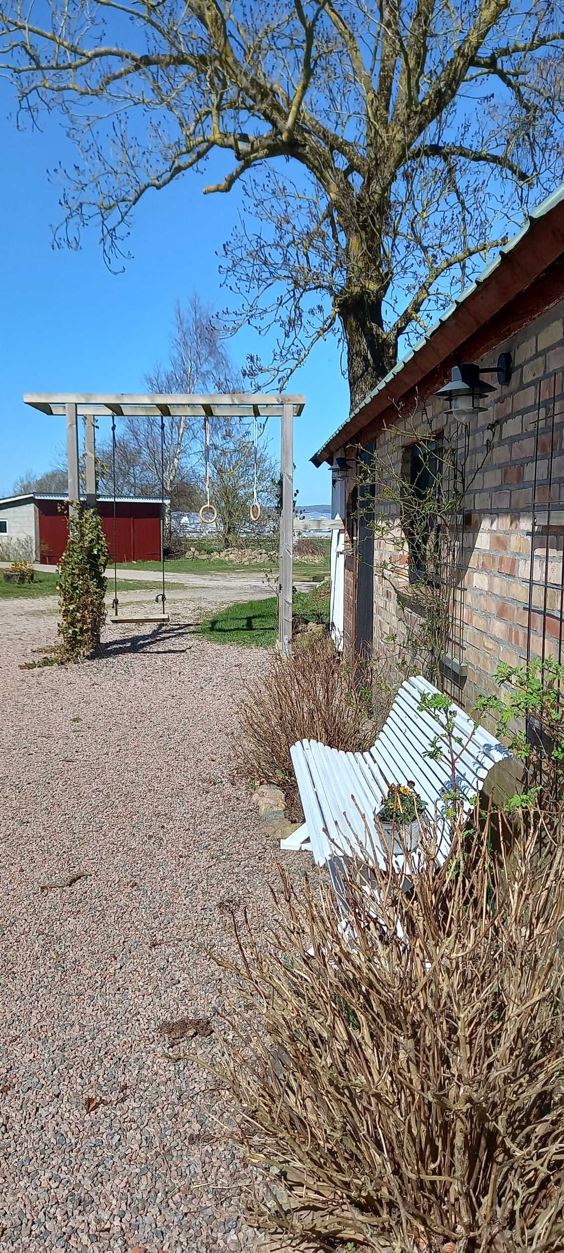 A gravel pathway leads to a light-colored bench positioned beside a brick wall. A swing set is visible in the background, framed by trees and open fields under a clear blue sky.