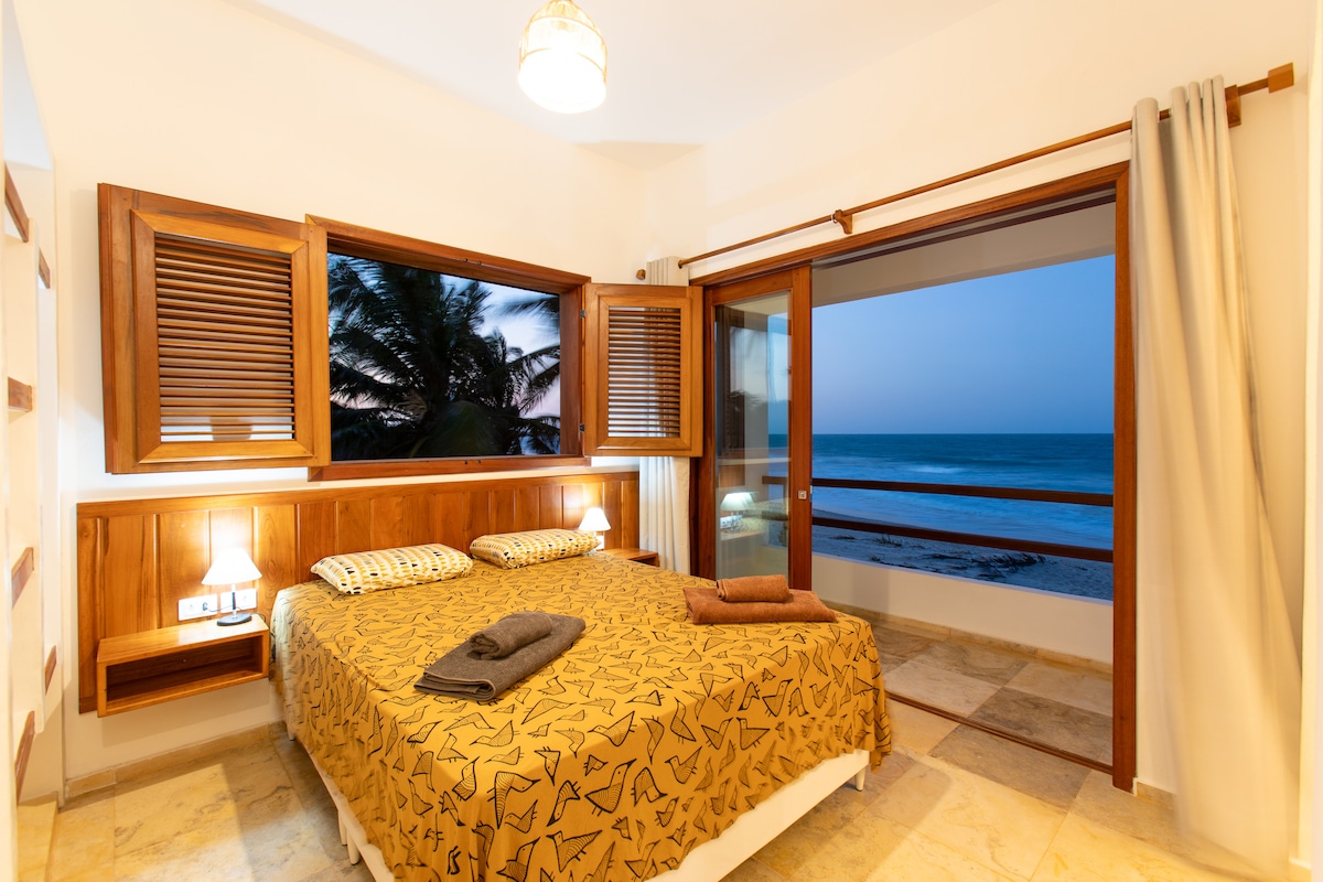 A bright bedroom features large windows that open to breathtaking ocean views. Two queen-size beds are neatly arranged with soft linens and towels displayed. Wooden accents and warm lighting create a cozy ambiance, while the gentle sound of waves can be heard nearby.