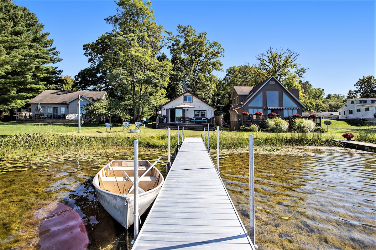A wooden dock extends over the water, leading to a lake house surrounded by grassy areas and trees. A small rowboat is moored alongside the dock. The tranquil scene is bathed in natural light, highlighting the home's large windows and inviting exterior.