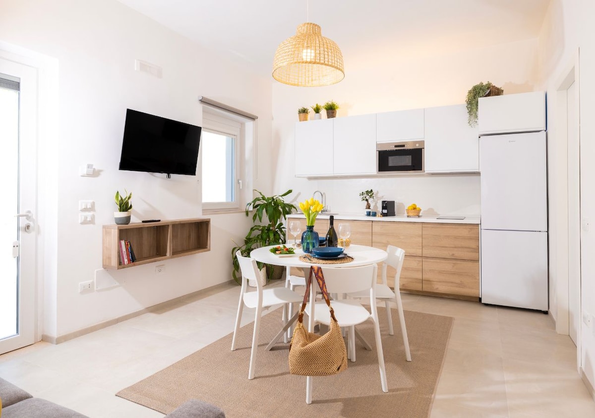 An open-plan kitchen and dining area displays modern white cabinetry and wood accents. A round table is set for four, surrounded by white chairs. A large window allows natural light, and a television is mounted on the wall. Indoor plants add a touch of greenery.