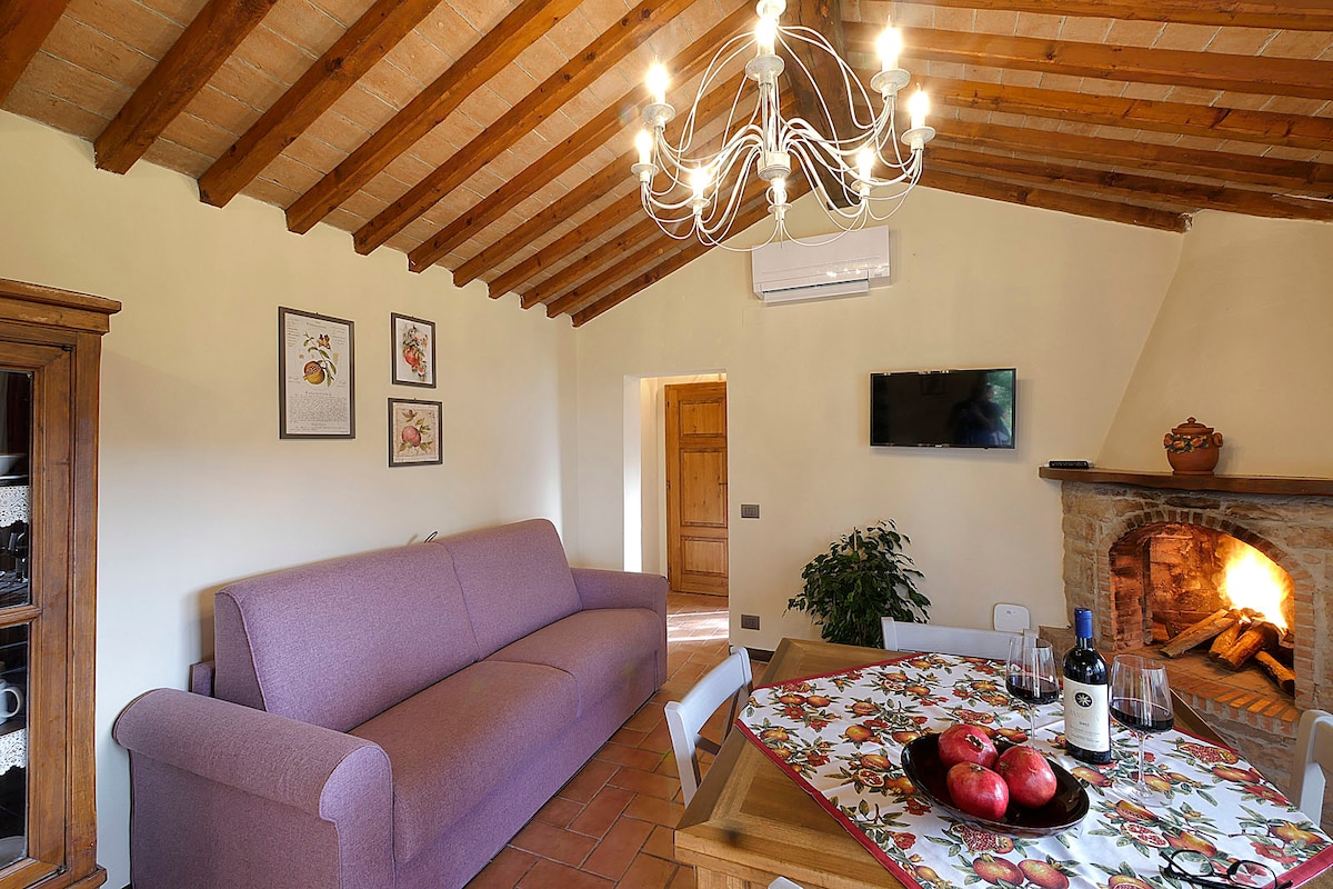 The living area features exposed wooden beams and a warm color palette. A comfortable sofa is positioned near a rustic fireplace, complemented by a dining table set with a decorative tablecloth showcasing fresh fruit. A flat-screen TV is mounted on the wall, enhancing the space's functionality.