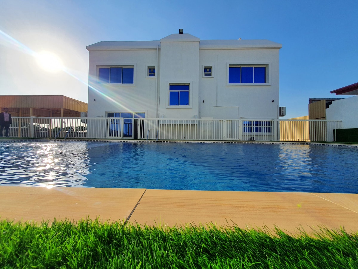 The villa features a modern exterior with large windows reflecting the sunlight. A private swimming pool is positioned in the foreground, bordered by neatly trimmed grass. Lounge chairs and shaded areas are visible in the background, providing spaces for relaxation.