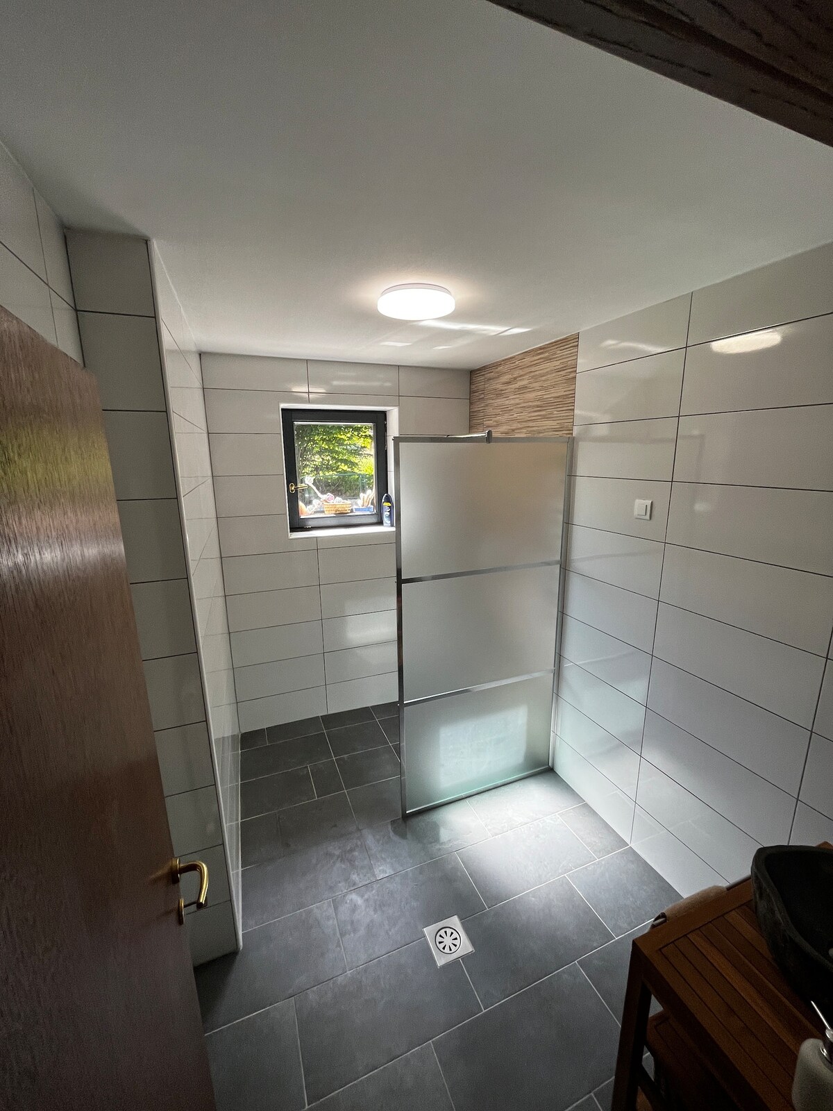 A modern bathroom is presented with a spacious shower enclosure featuring a frosted glass door. Natural light enters from a window, illuminating the sleek tile walls and floor. A small wooden chair and a drainage grate add practical elements to the streamlined design.