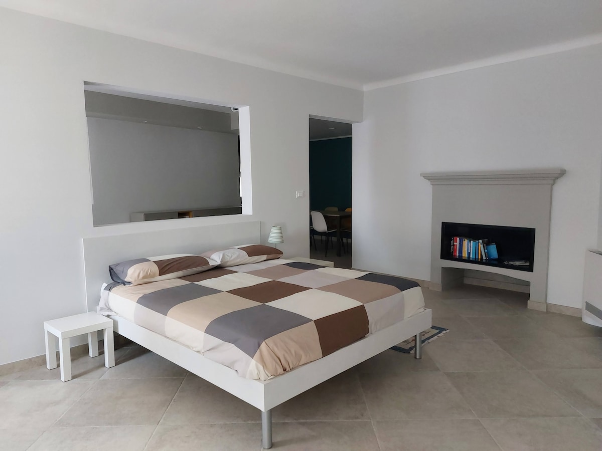 A spacious bedroom features a large bed with a checkered duvet in neutral tones. A bedside table holds a lamp, and a modern fireplace adds warmth to the room. Natural light enters through large windows, enhancing the airy atmosphere.