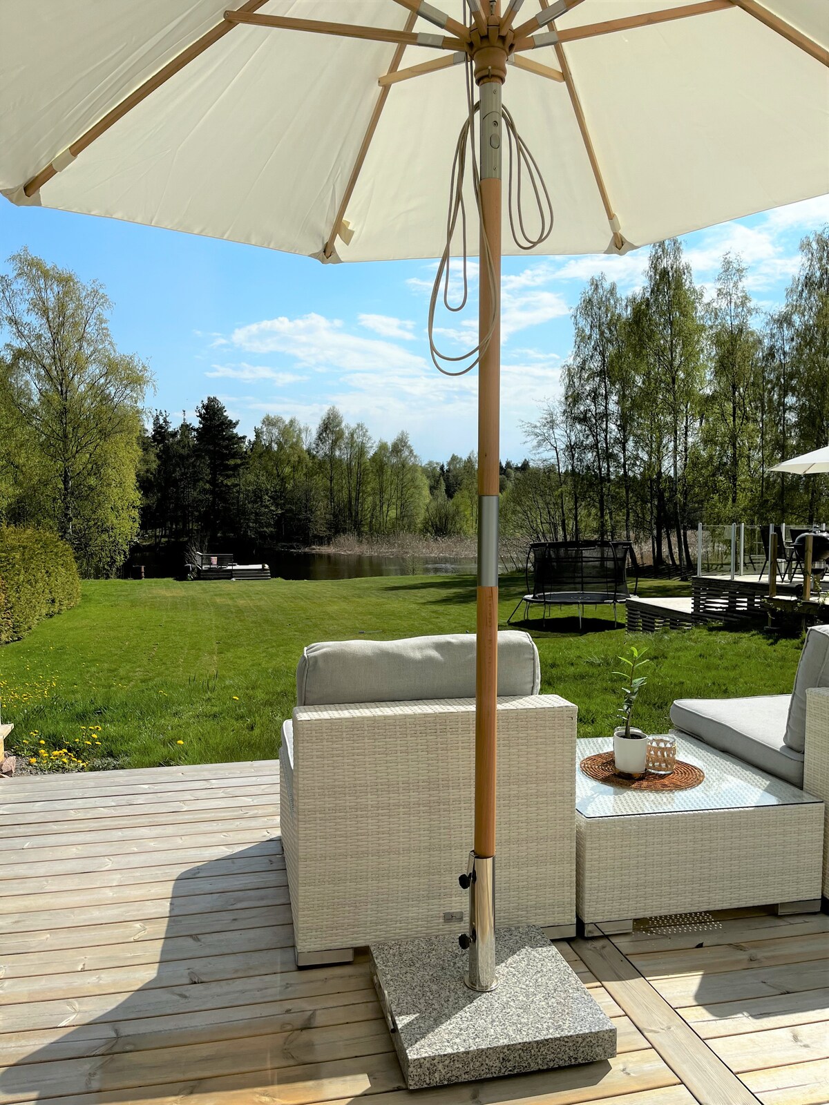 A shaded outdoor seating area features light-colored wicker chairs with plush cushions. A large umbrella stands over a small table adorned with a decorative tray. Lush green grass extends to the water's edge, surrounded by trees under a clear blue sky.