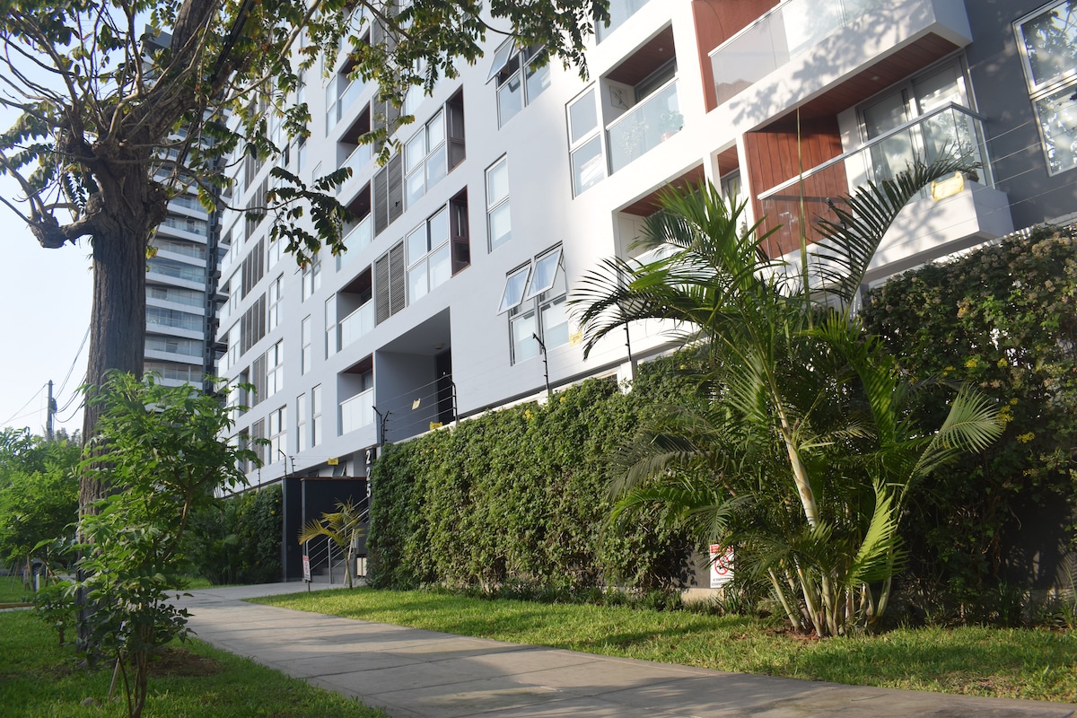 A modern apartment building is framed by lush greenery and well-maintained landscaping. A wide, paved walkway leads to the entrance, bordered by shrubs and trees. The facade features a mix of light and dark materials, with large windows reflecting the surrounding nature.