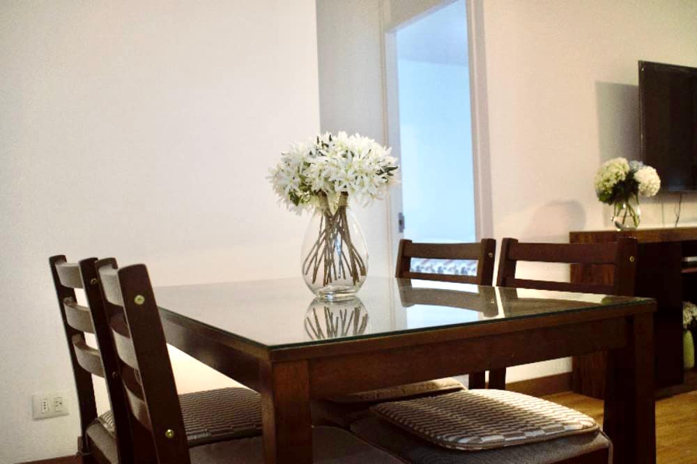 A wooden dining table is centered in the room, surrounded by four cushioned chairs. A vase of white flowers sits on the table's glass surface, adding a touch of nature. A door frame is visible in the background, leading to another area of the apartment.
