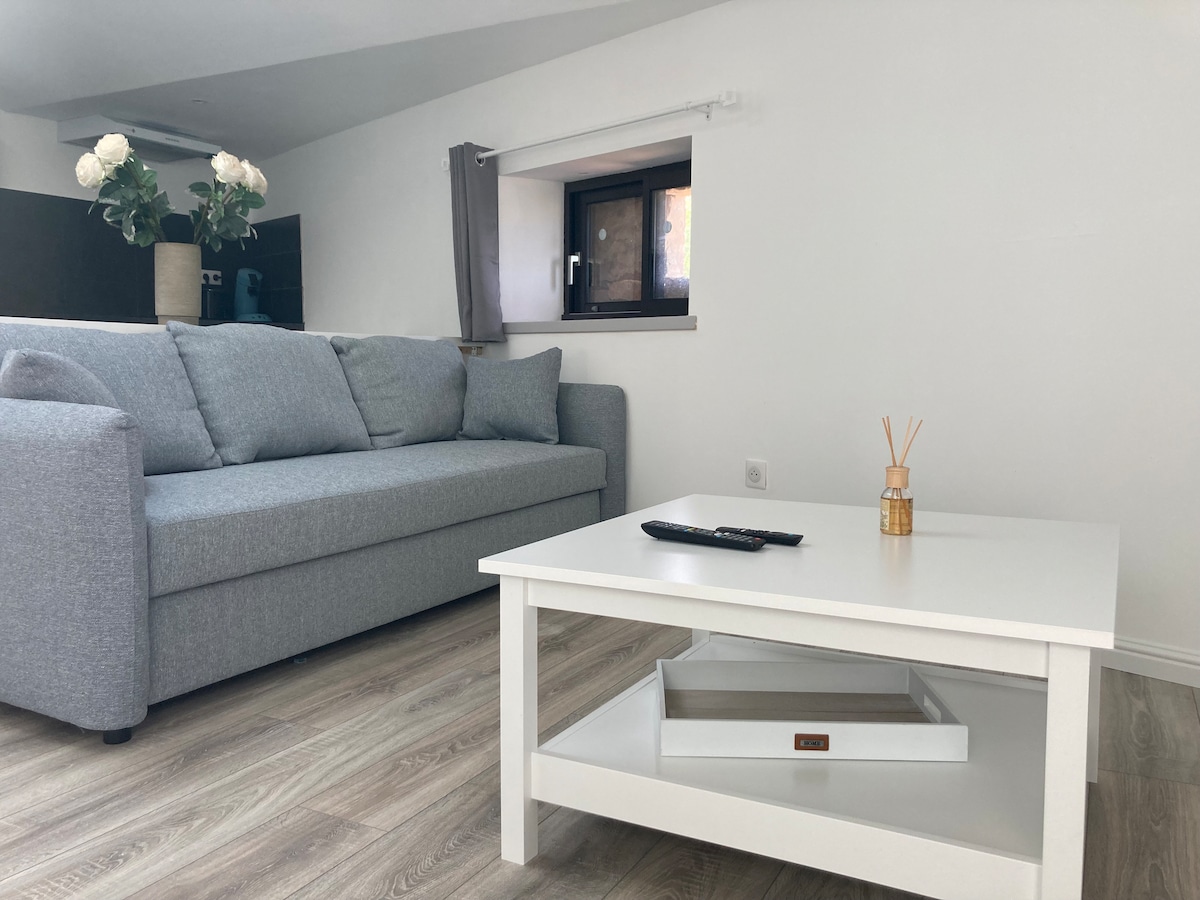 A bright living area features a light grey sofa complemented by a white coffee table. The coffee table includes a decorative tray, a remote control, and a reed diffuser. Natural light enters through the window, enhancing the neutral decor and modern finishes.