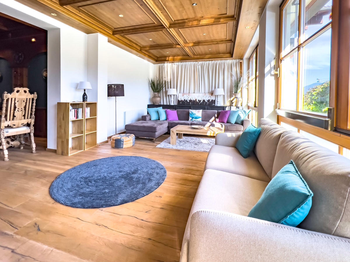 A spacious living area features large windows allowing natural light to fill the room. Comfortable seating is arranged around a central coffee table, with decorative cushions adding a pop of color. A warm wooden ceiling enhances the cozy ambiance, while a soft rug anchors the seating space.