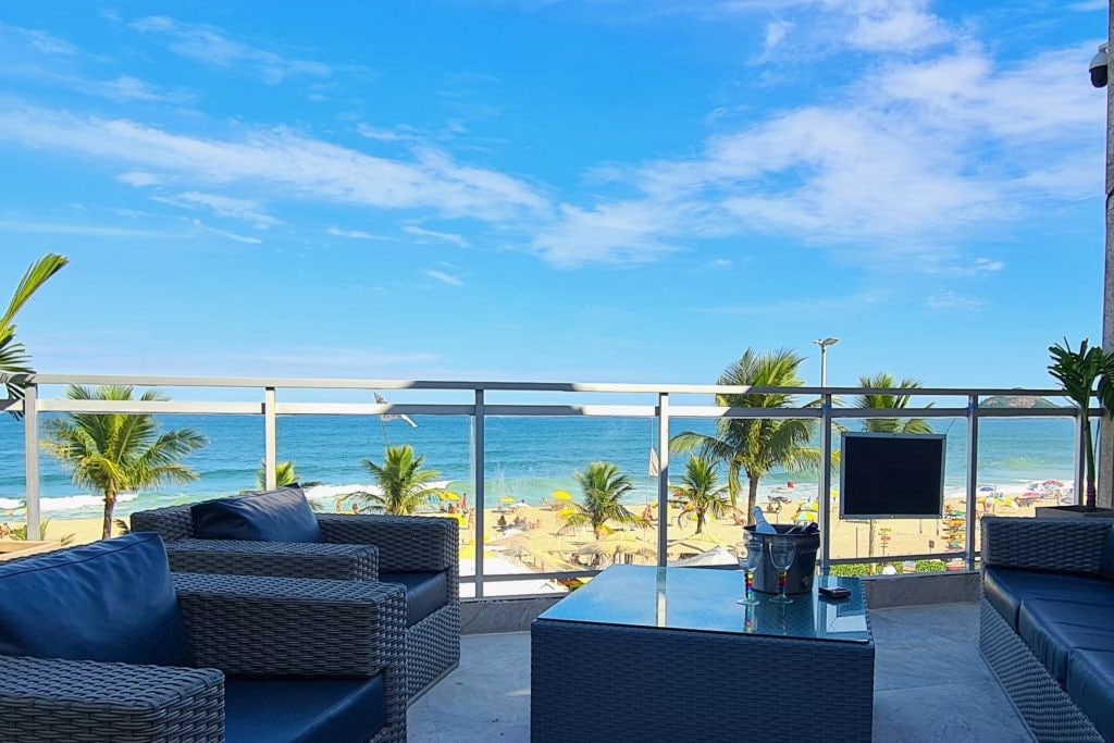A spacious outdoor lounge is presented, featuring comfortable seating and a glass-top table. The view showcases a pristine beach with soft waves and palm trees, creating a serene backdrop against the bright blue sky.