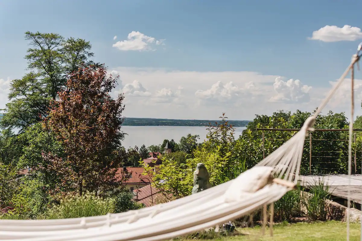 A hammock is positioned in a lush garden, framing a panoramic view of the Ammersee. Surrounding green trees and colorful foliage enhance the serene atmosphere, while the lake glimmers in the distance under a clear blue sky.