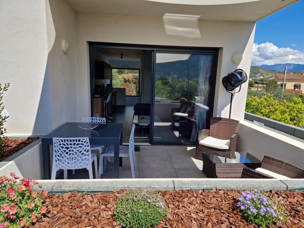 A shaded terrace features a dining table surrounded by white chairs and a seating area with wicker furniture. Lush greenery can be seen in the background, and the spacious area is illuminated by natural light from the large sliding doors that connect to the interior.