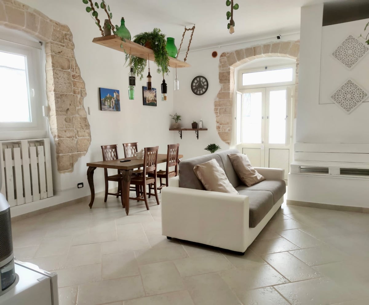 A spacious living area features a comfortable white sofa opposite a wooden dining table with four chairs. Natural light brightens the space through large windows framed by stone accents. Decorative plants and a wall clock add a touch of charm to the ambiance.