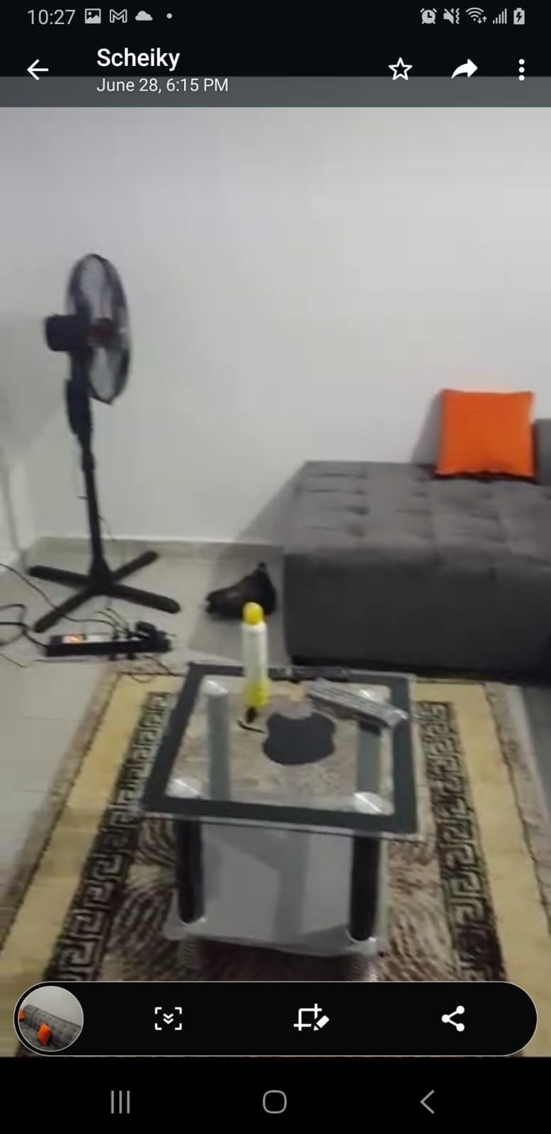 A cozy living area features a gray sectional sofa with orange accent pillows. A small glass coffee table sits on a patterned rug, accompanied by a candle and a decorative item. A standing fan is positioned nearby, contributing to the airy environment.