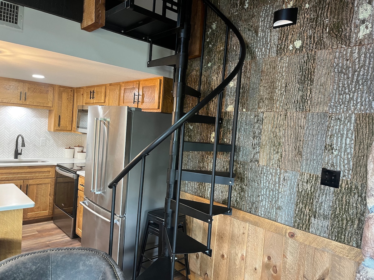 A modern kitchen is visible adjacent to a spiral staircase with a sleek black design. The wooden cabinets and stainless steel appliances complement the rustic wall featuring natural wood textures. Soft lighting enhances the inviting atmosphere of the space.