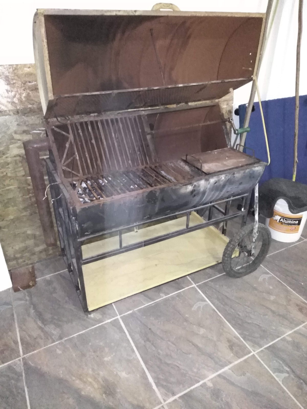 A charcoal grill is positioned on a smooth tiled surface. The grill features a large cooking area with a raised lid and a wheeled base for mobility. The surroundings include a partially visible wall and storage area. Various tools are organized nearby.