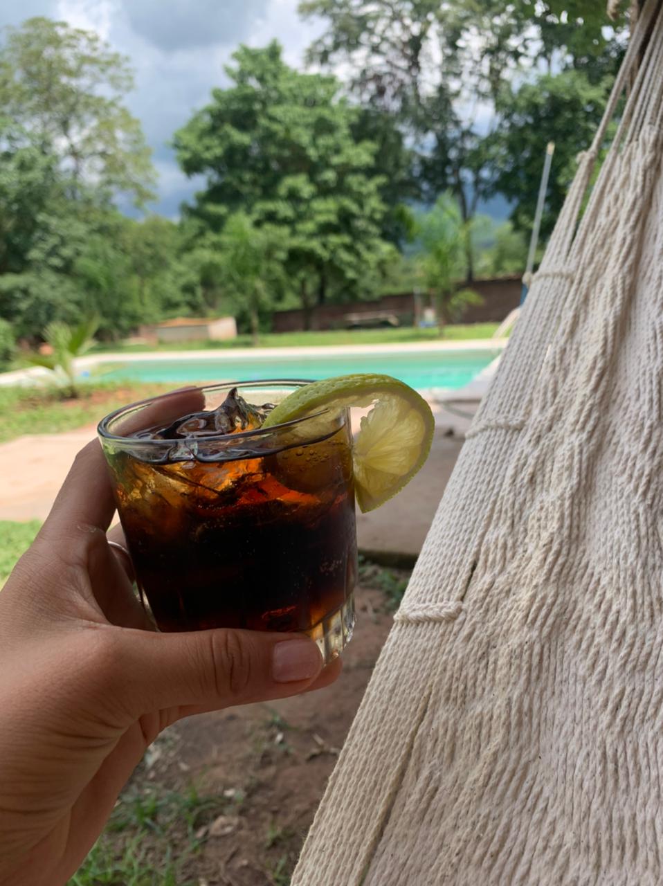 A close-up of a refreshing beverage in a glass is captured against a backdrop of lush green trees and a shimmering pool. A slice of lime rests on the rim, while a hammock is visible nearby, suggesting a relaxed outdoor setting.