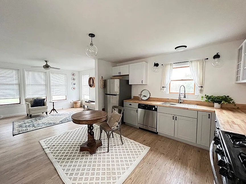 A modern kitchen area features light-colored cabinetry and a wooden countertop, complemented by stainless steel appliances. A round wooden dining table with a single chair is positioned on a patterned rug. Large windows allow natural light to illuminate the space, creating an airy atmosphere.