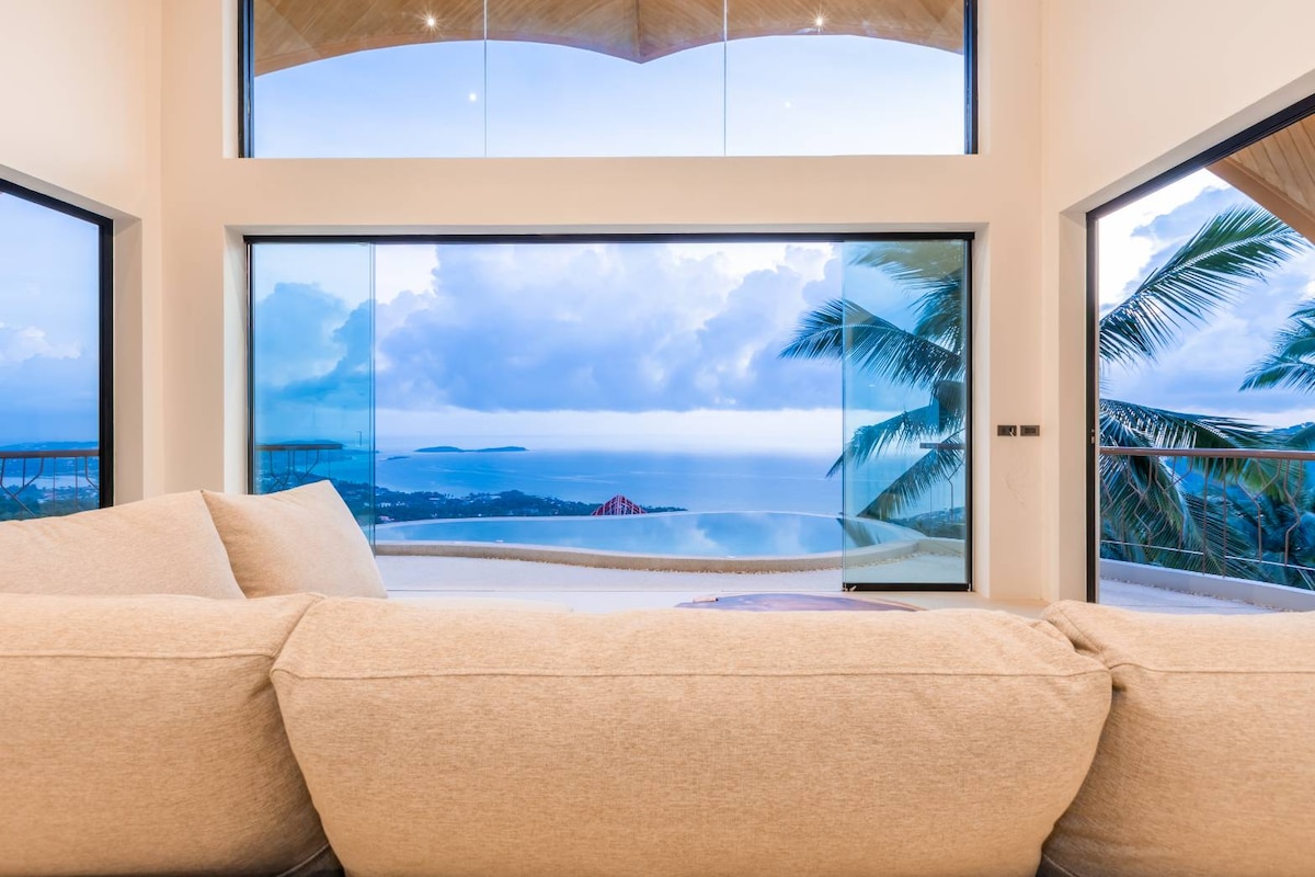A spacious living area features a large, soft sofa facing large floor-to-ceiling windows offering unobstructed views of the sea and distant islands. Natural light fills the space, highlighting the modern design and inviting atmosphere.