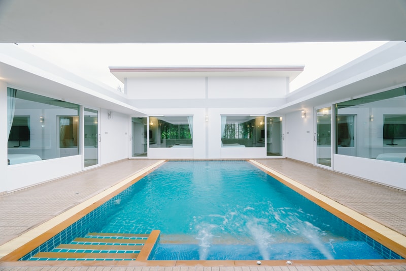 A swimming pool is centered in an open courtyard, surrounded by modern glass doors that provide views into the adjacent rooms. The pool features a gentle blue hue with steps leading into the water, complemented by a light-colored deck and a serene ambiance.