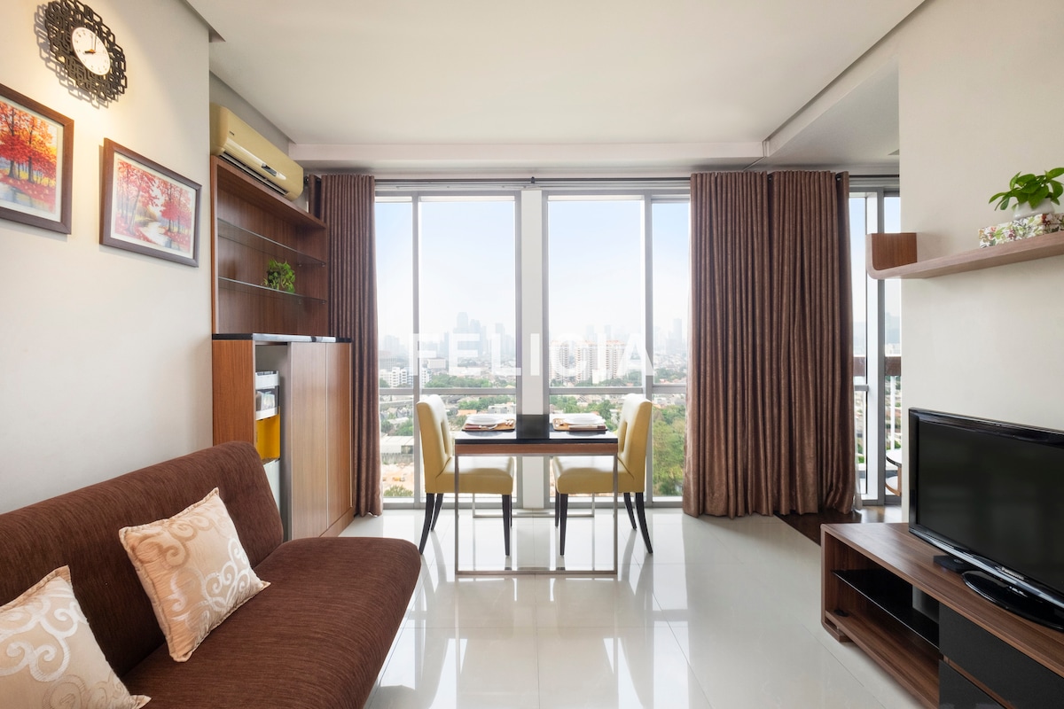 A modern living area features a comfortable sofa bed, a dining table for two, and a flat-screen TV. Large windows provide a view of the cityscape, with natural light illuminating the space. Curtains frame the windows, enhancing the cozy ambiance.