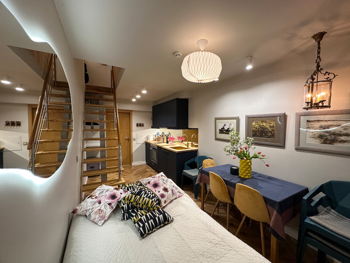 A cozy space with a spacious bed adorned with patterned pillows is visible. A dining area with a dark table and yellow chairs is present, alongside a staircase leading to an upper level. Soft lighting highlights the kitchen area, creating a warm ambiance.
