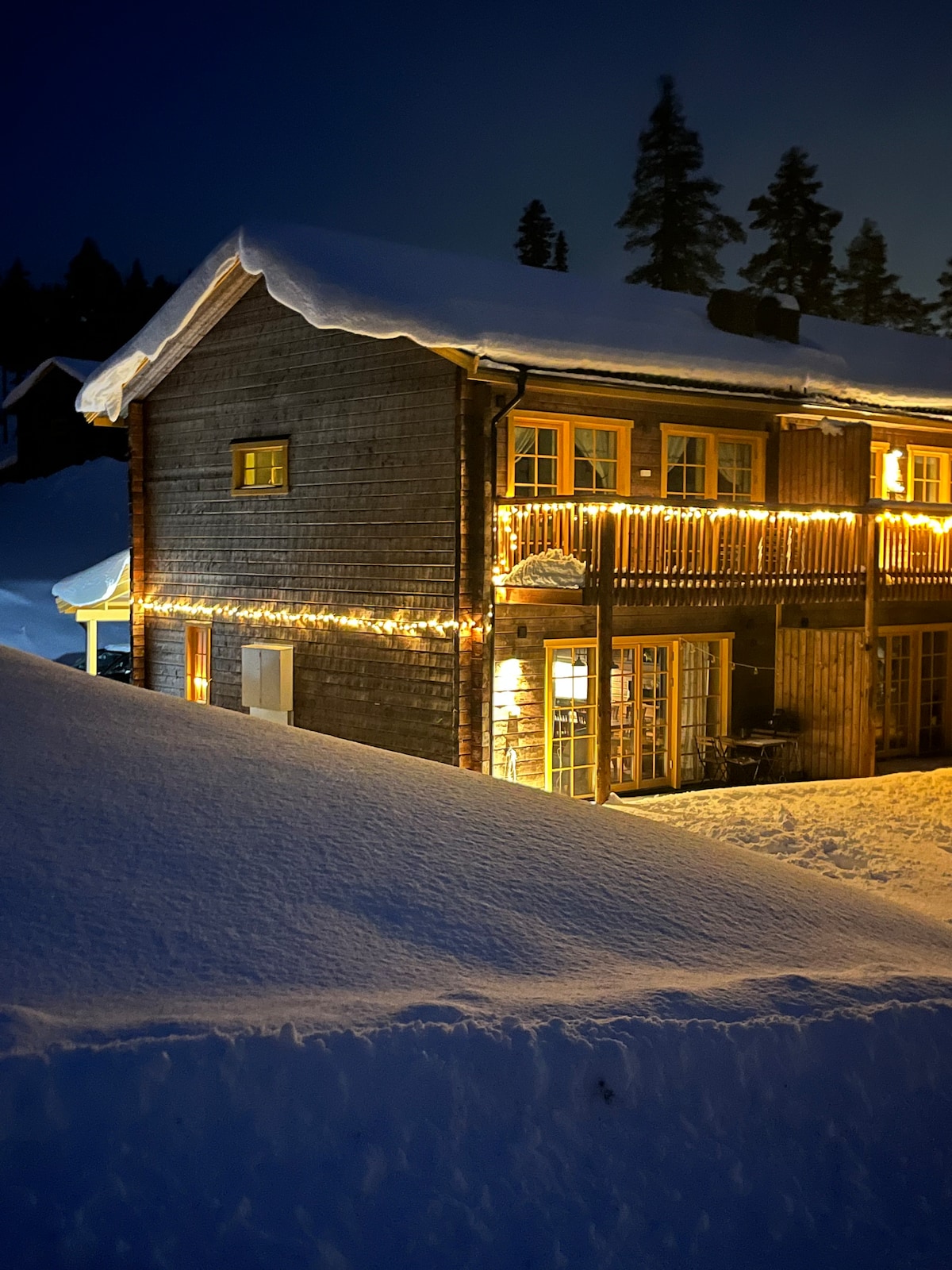 The wooden exterior of the residence is adorned with soft lighting, creating a warm glow against the snowy landscape. Snow blankets the ground, highlighting the building's charming architecture. Trees are visible in the background, adding to the serene winter atmosphere.