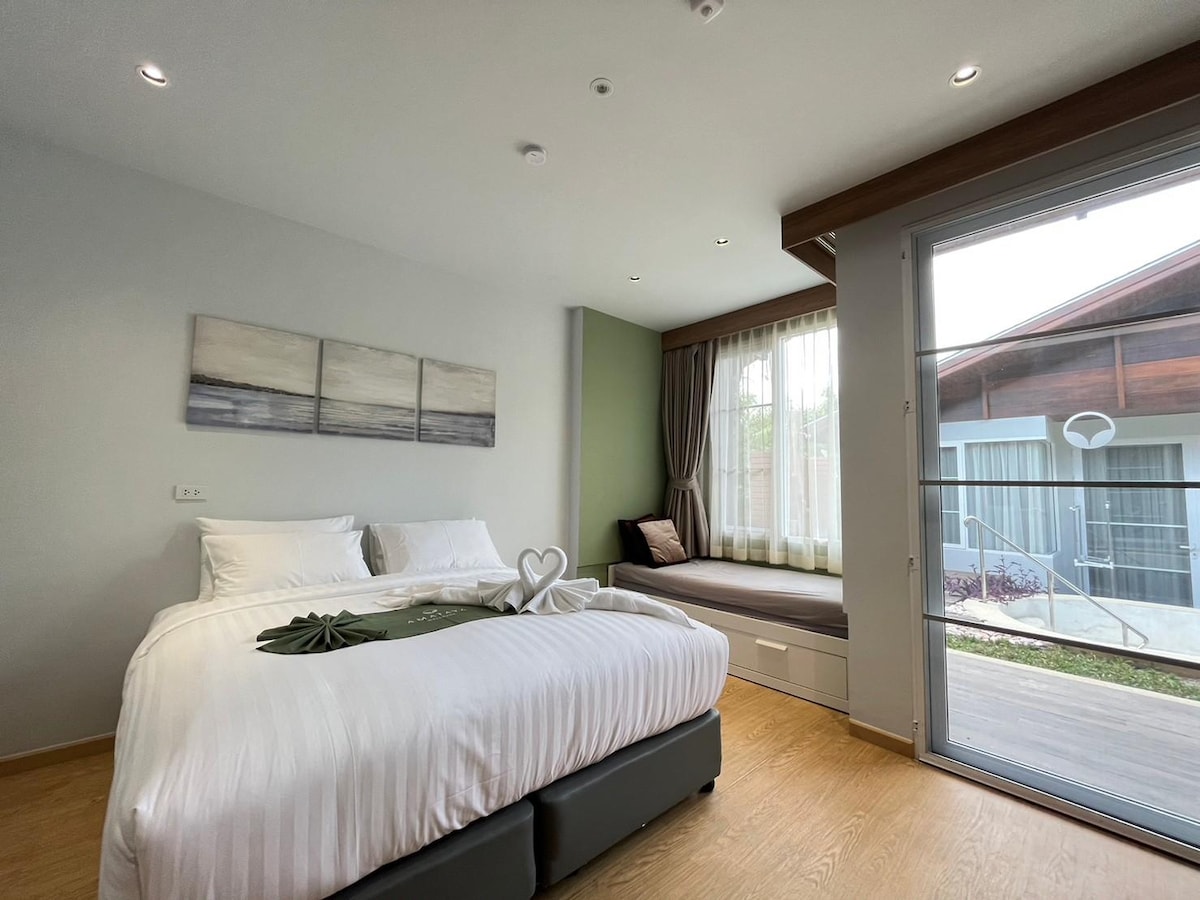 The room features a spacious bed with crisp linens and decorative accents, positioned next to large glass doors that provide natural light. A cozy seating area is nearby, offering a view of the outdoor space.
