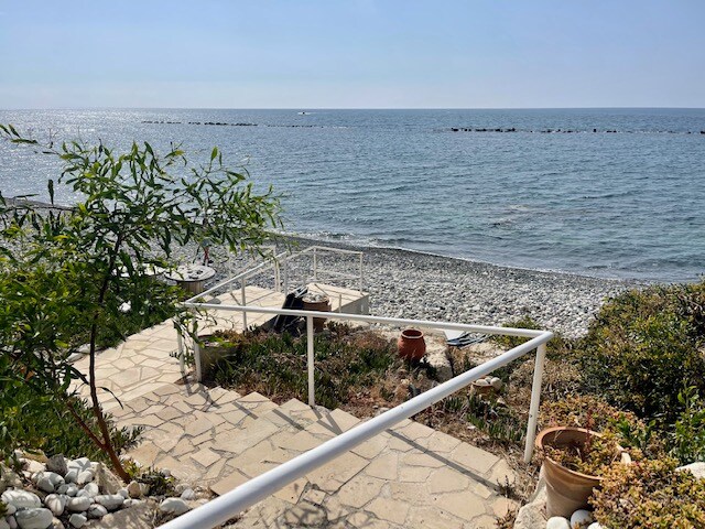 A pathway descends toward the beach, surrounded by greenery and decorative pots. The smooth stones and pebbles form a natural shore alongside the calm, blue water. In the distance, a few boats can be seen on the horizon under a clear sky.