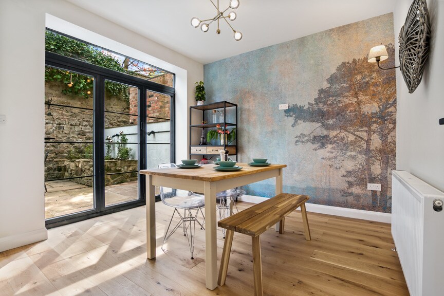 A dining area is displayed with a wooden table and bench seating, accompanied by carefully placed tableware. A feature wall with an artistic design is visible, enhancing the ambiance. Bi-fold doors open to a private outdoor space, allowing natural light to illuminate the room.