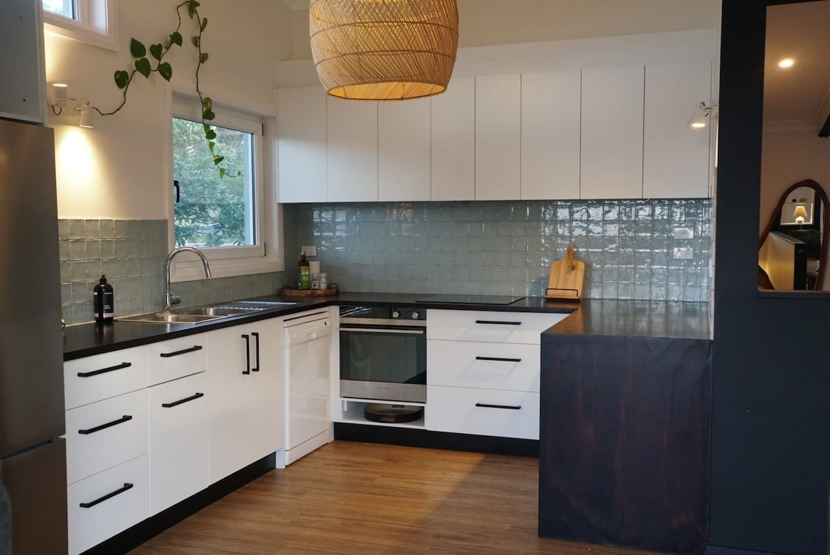 The modern kitchen features white cabinetry with sleek black hardware and a dark countertop. Soft blue tiles cover the walls, while natural light enters through a nearby window. Equipped with an induction cooktop, oven, dishwasher, and sink, the space combines functionality with a clean design.
