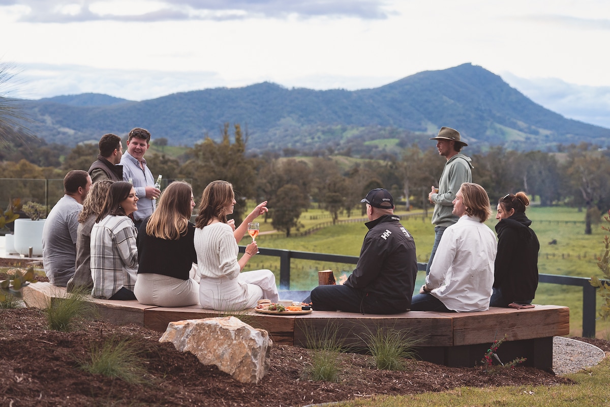 A group of individuals is seated on a wooden bench, enjoying drinks and snacks. The backdrop features rolling hills and mountains, creating a natural landscape. The scene captures a relaxed outdoor gathering, framed by greenery and scattered rocks.