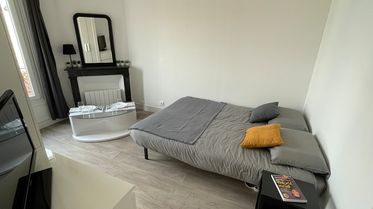 The room features a spacious bed with a gray cover, accompanied by decorative pillows and a soft throw. A modern coffee table is positioned in front of the bed. A mirror and a flat-screen TV are visible against the neutral wall, enhancing the room's functionality.