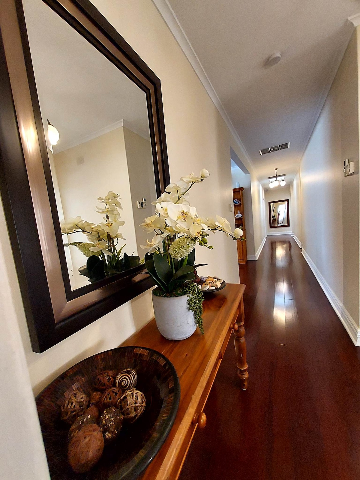 A long hallway features polished wooden flooring and soft neutral walls. A mirror with a dark frame is positioned above a wooden side table adorned with a decorative bowl and a potted orchid. The distant end of the hallway leads to additional space lit by natural light.
