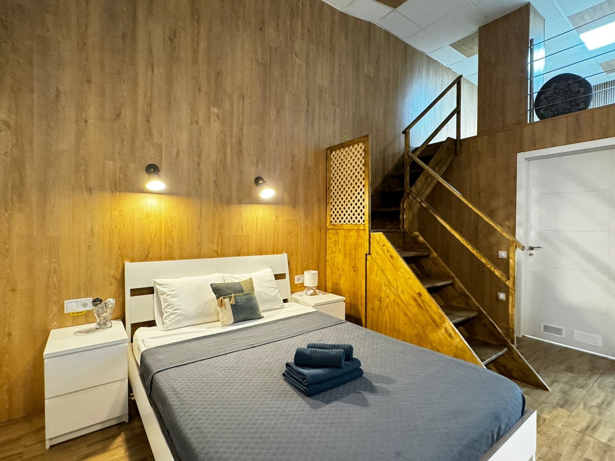 A cozy bedroom features warm wooden walls and a spacious bed with neatly arranged towels. Two light fixtures provide illumination alongside a nightstand. A staircase leads to an upper level, enhancing the room's character and openness.