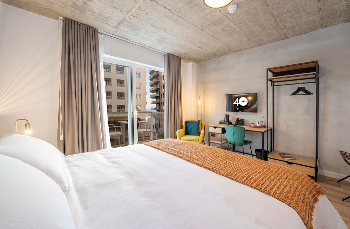 A spacious guestroom features a large bed with a textured throw, complemented by contemporary furnishings. A desk and chair are positioned near large windows, allowing natural light to fill the room. A sleek TV and clothing rack are also visible, creating a modern ambiance.