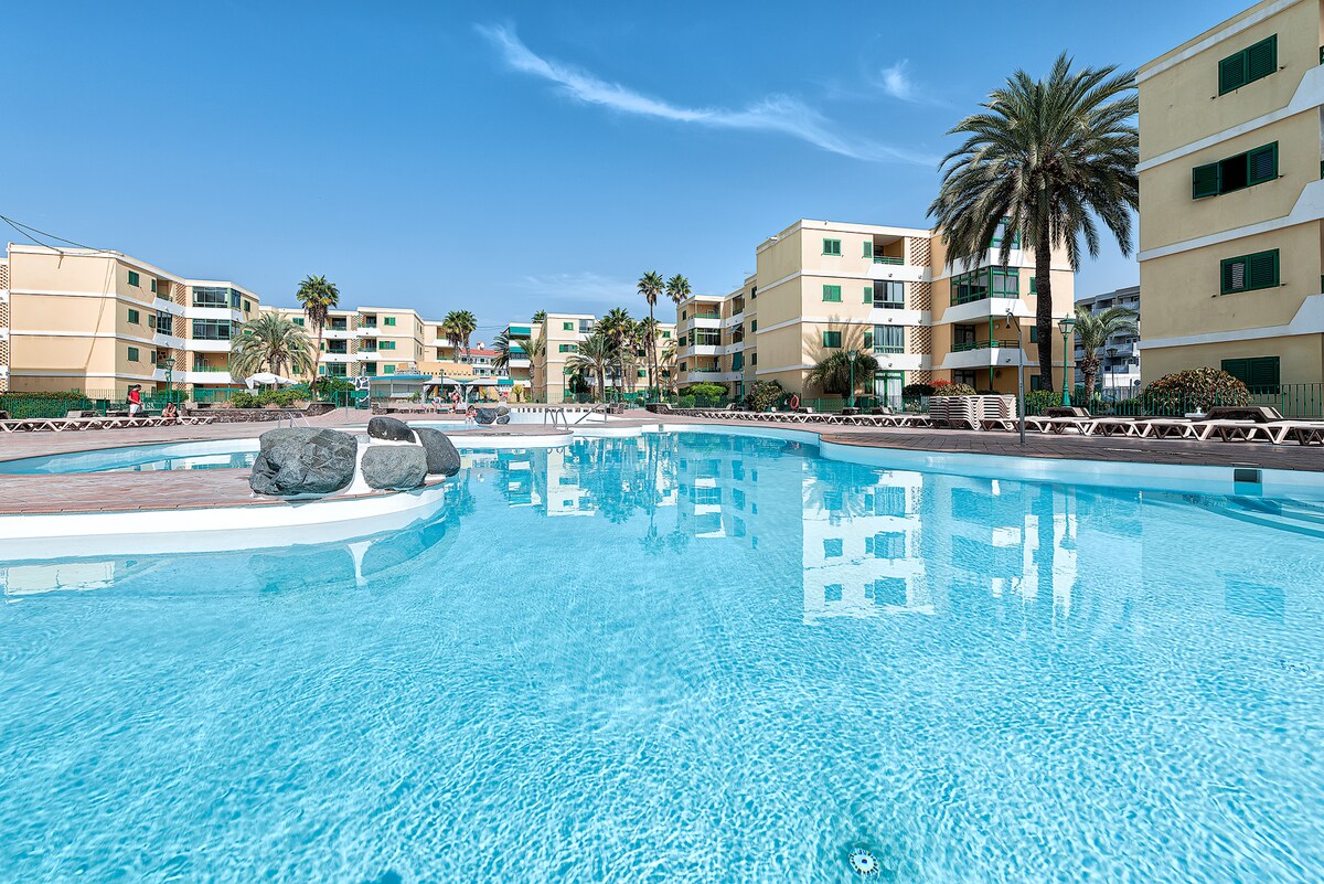 A large outdoor pool is surrounded by palm trees and lounge chairs. The clear blue water reflects the sunny sky. Multiple buildings with light-colored facades are visible in the background, creating an inviting atmosphere for relaxation.