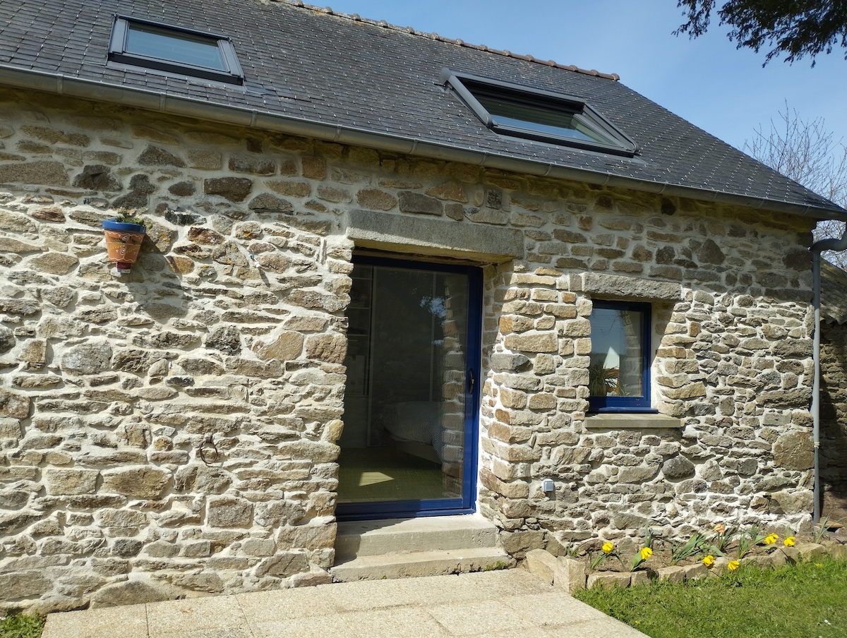 The exterior of the accommodation features beautifully textured stone walls, complemented by blue window frames and a matching door. Sunlight aligns with the entrance, showcasing well-maintained steps that lead to a small patio area, bordered by blooming flowers and greenery.