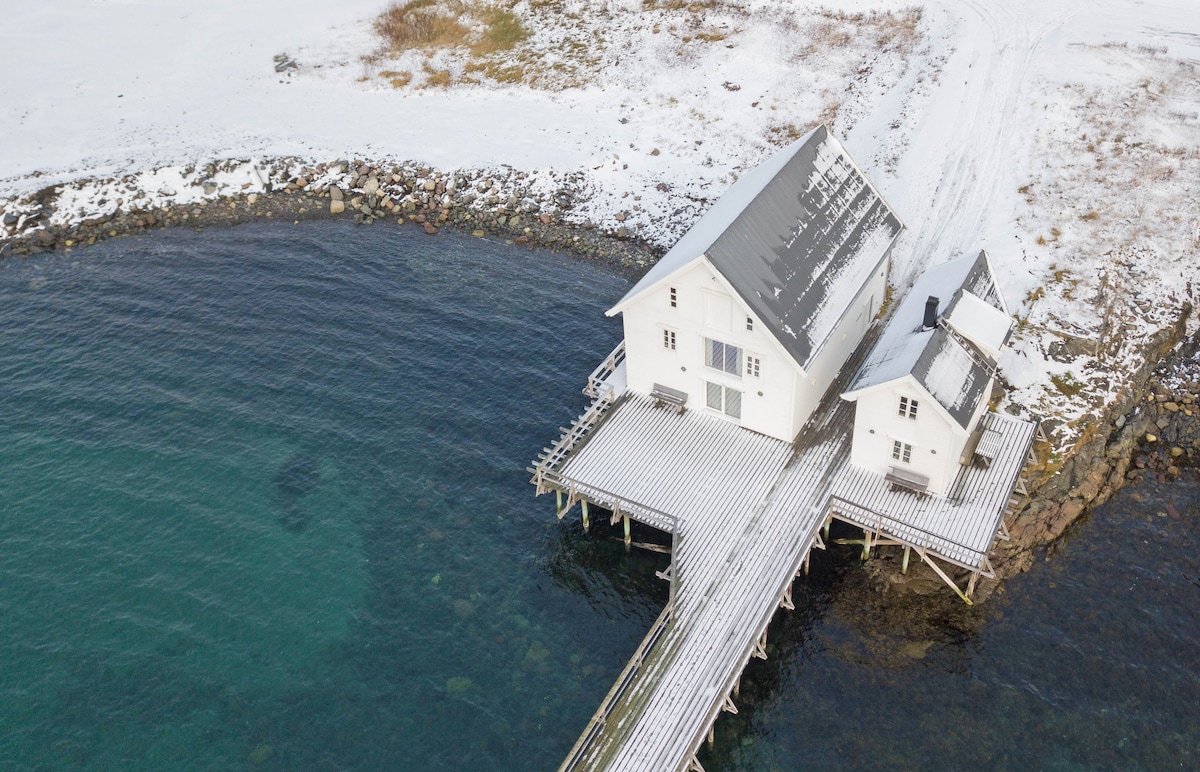 Two modern stilt apartments are situated over the calm, clear waters of the Barents Sea, connected by a wooden walkway. The exterior is characterized by white walls and a sloped roof, with patches of snow covering the surrounding landscape.
